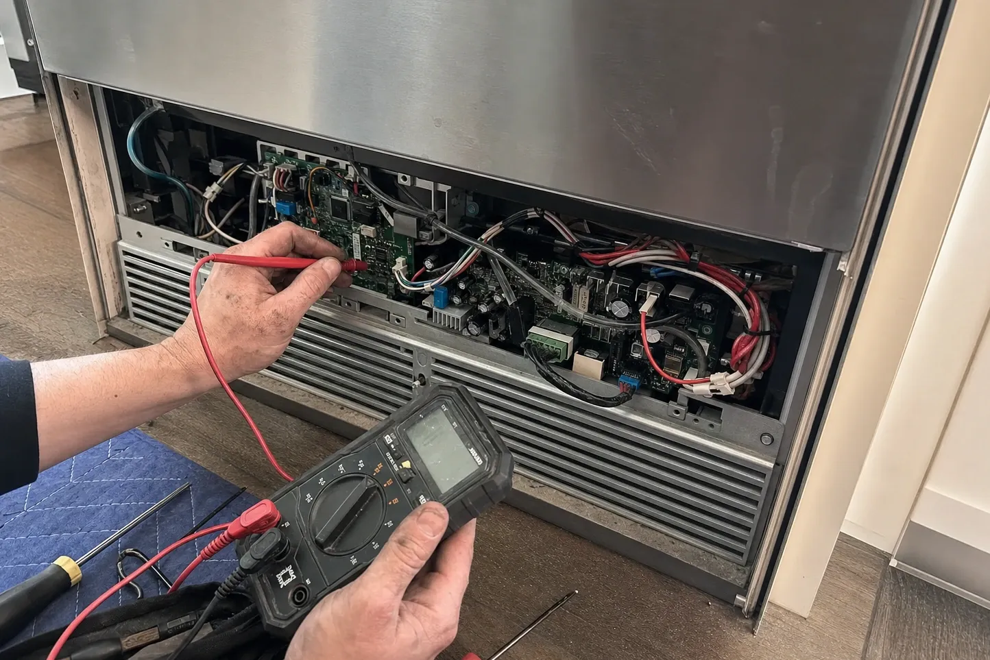 Hands with a multimeter probing a bank of exposed control boards behind the upper grille of a stainless built-in refrigerator