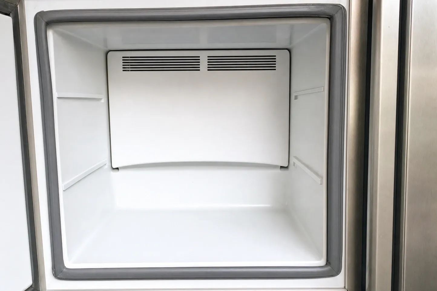 An empty freezer compartment framed by its grey door gasket, the rear panel carrying two banks of vent louvres above bare shelf ledges